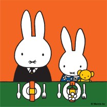 Two white rabbits sat eating food at the table with two plates in front of them and the smaller rabbit holding a teddy bear